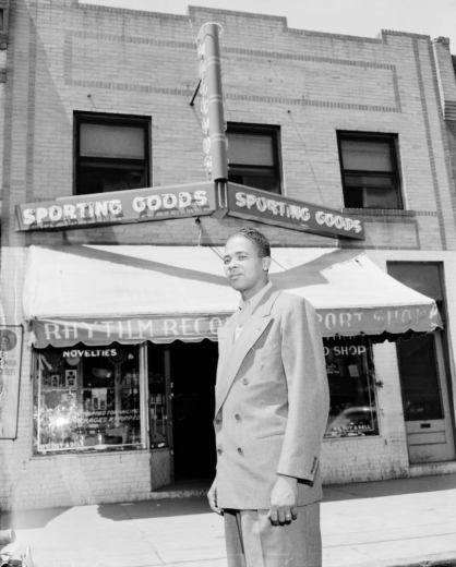 Leroy Smith, an African American, stands in front of Rhythm Records Sport Shop on 26th and Welton Street in the Five Points neighborhood of Denver, Colorado.  He wears a double breasted suit.  The store has an awning above the front windows and a neon sign that reads: "Sporting Goods" and "Records."
