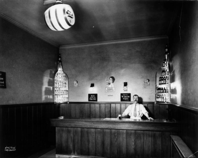 Interior view of the Tivoli Union Brewery tasting room in Denver, Colorado; Harry Pfalzgraf poses at the bar. Signs read: "Beer Issued on Chips Only," and "Positively No Loafing Allowed."