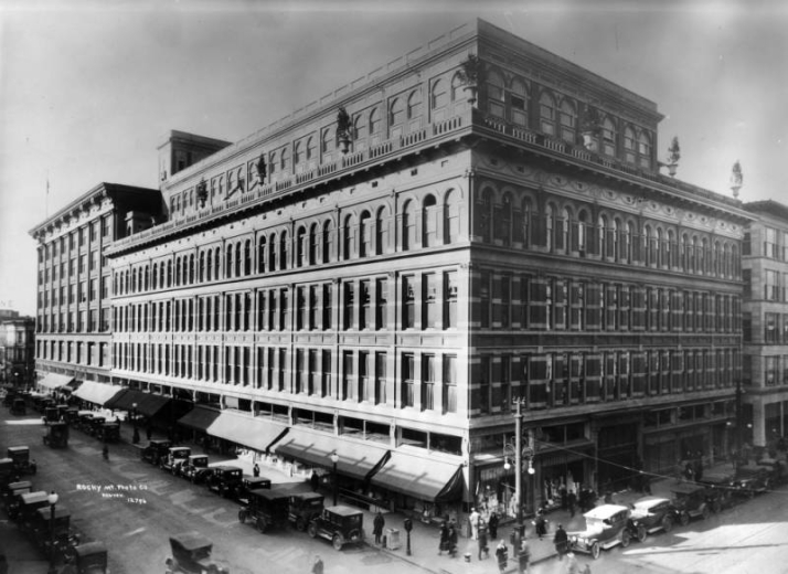 View of the Denver Dry Goods Store, at 16th (Sixteenth) and California Streets, in downtown Denver, Colorado; shows a brick and stone building with a roof terrace and urns. Pedestrian and automobile traffic fill the streets.