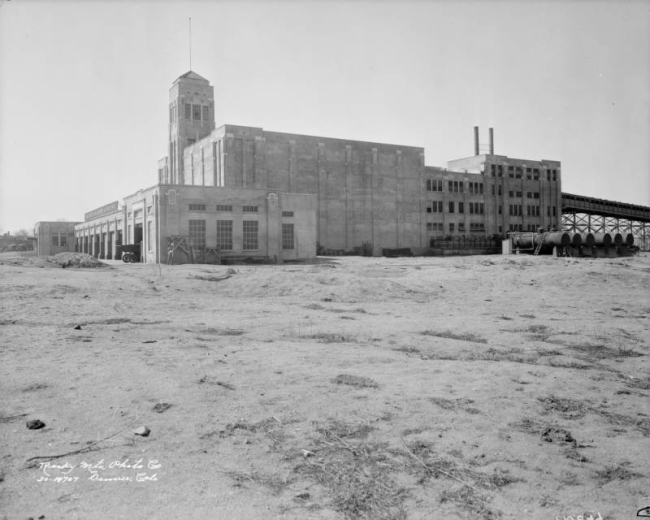 View of the Blayney-Murphy slaughterhouse at 45th (forty fifth) and Gilpin Streets in the Elyria Nieghborhood of Denver, Colorado; shows a concrete building with truck bays, a tower, and smokestacks.