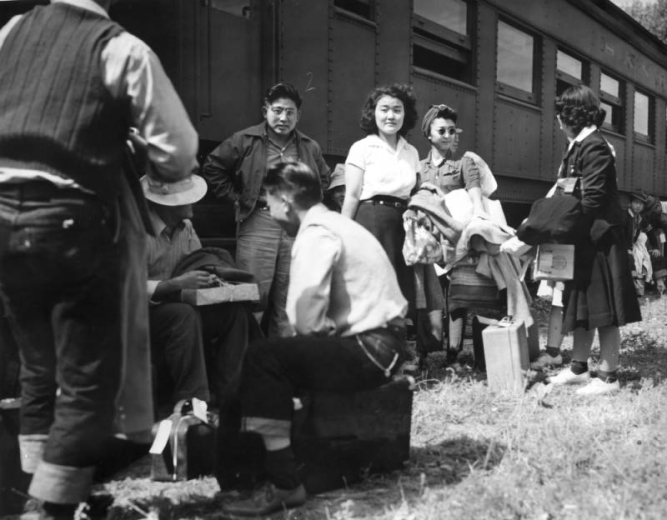 Japanese evacuees stand or sit with their suitcases and belongings in front of a Santa Fe and Topeka passenger train car. The men and women wait for the bus ride to Camp Amache, Granada Relocation Center, southeastern Colorado.