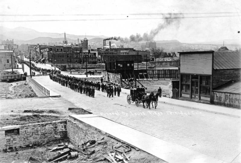 Hundreds of coal miners march in a funeral procession for Ludlow Massacre victim Louis Tikas, on North Commercial Street in Trinidad, Las Animas County, Colorado during the UMW labor strike against CF&I. Business signs read: "Laundry", "Trinidad Hotel", "[New Met]ropolitan Hotel", "Wrigley's Spearment", "Colorado [S]upply Co.", and "The Pierc[e.]"