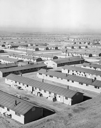Overview of the Granada Relocation Center, Camp Amache, Prowers County, southeastern Colorado, shows rows of prefabricated army-style barracks (120 x 20 feet.) Twelve of these one-story side gable barracks with tar-paper roofs made a block which had its own recreation hall, laundry, and mess hall. Wooden storage boxes, benches, palettes, and laundry lines show around buildings. Prairie and bare trees are in the distance.
