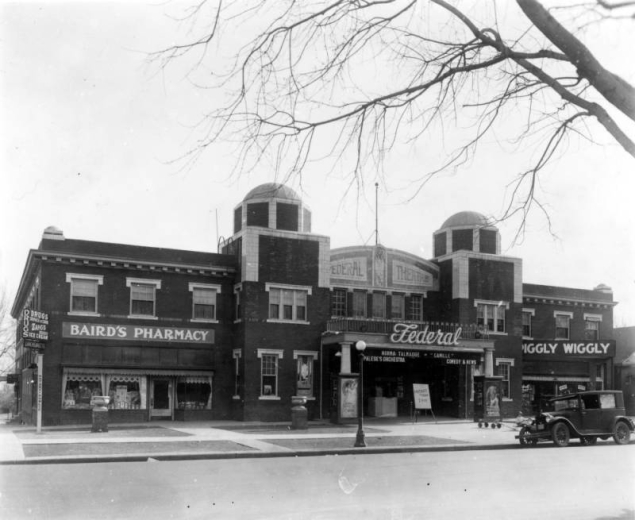 View of the Federal Theater near 38th (Thirty-eighth) Avenue and Federal Boulevard in the Sunnyside neighborhood of Denver, Colorado. The two-story brick building has towers topped with domes. Signs read: "Baird's Pharmacy," "Drugs Service at Curb Zang's Delicious Ice Cream," "Luncheonette," "Norma Talmadge Camille Palese's Orchestra Comedy & News," and "Piggly Wiggly."