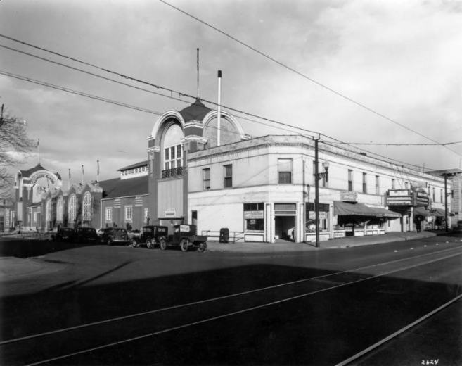 View of Mammoth Gardens at Colfax Avenue and Clarkson Street in the North Capitol Hill neighborhood of Denver, Colorado. The two-story building has Romanesque arched windows and domed towers with balconies. Automobiles and pick-up trucks are parked nearby. Signs read: "Used Cars," "Mammoth Garden," "Roller Skating," "Archer Brothers Pascal Celery Shippers," "Mammoth Garden Market," and "Hotel."