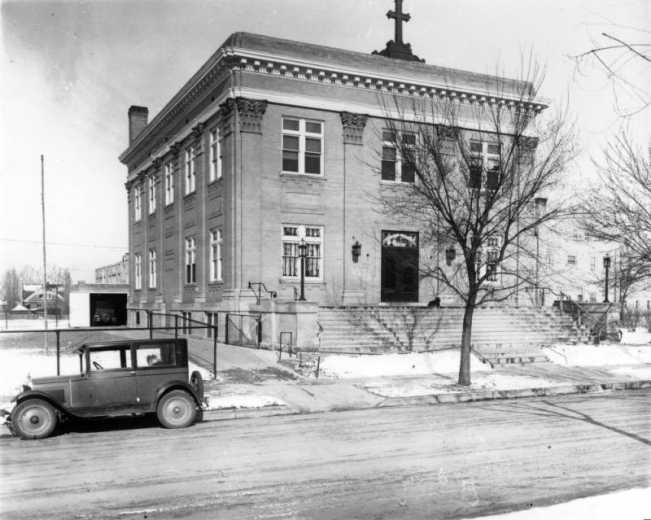 View of the Blessed Sacrament Church at Montview Boulevard and Elm Street in the South Park Hill neighborhood of Denver, Colorado. The two-story brick building (built 1913) has decorative trim and lintels. An automobile is parked nearby.