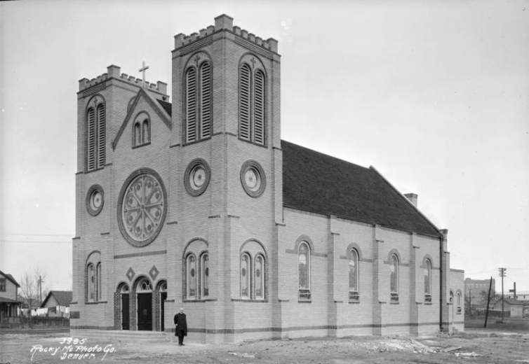 A priest stands near the Holy Rosary Church at 4695 Pearl Street in the Globeville neighborhood of Denver, Colorado. The brick building has bell towers, an arched entryway, and circular stained glass windows.