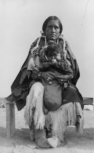 Outdoor portrait (sitting) of a Native American (Cheyenne) woman and child. The woman holds the child wrapped in a blanket and wears moccasins and a fringed dress and shawl.