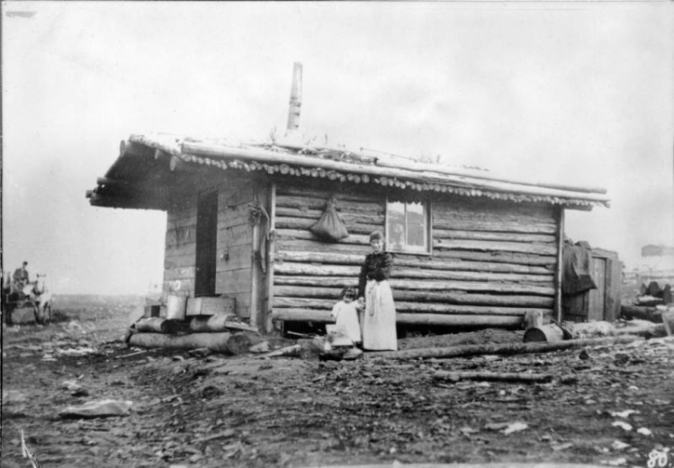 A woman and a young girl pose near a log cabin on a snow dusted landscape as a man driving a wagon approaches in the background, Colorado. The cabin has a plank front with timber walls and roof.