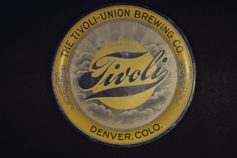 View of  a beer cap (ca.1901-1915) from the Tivoli-Union Brewery in Denver, Colorado. Shows a gold colored bottle cap with a central image of the sun and rays that radiate outward on a field of gray clouds. The words: "Trade Mark" and "Tivoli" are superimposed over the sun. The cap has a gold rim with Art-Nouveau designs and the words: "The Tivoli-Union Brewing Co., Denver, Colo."