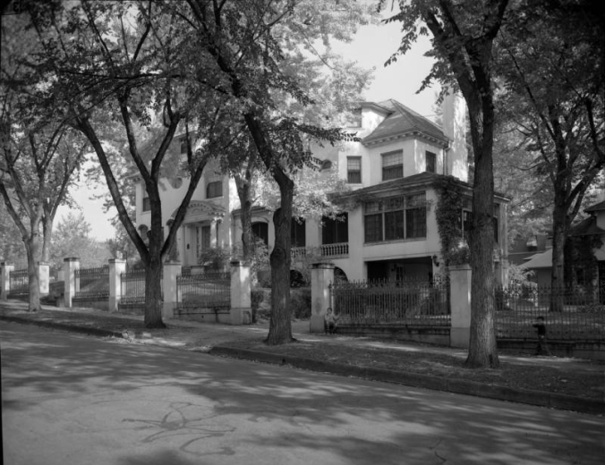 View of the Mitchell residence at 680 Clarkson Street in the Speer neighborhood of Denver, Colorado; features a portico, sun porch, and arcade. A boy sits by a wrought iron fence.