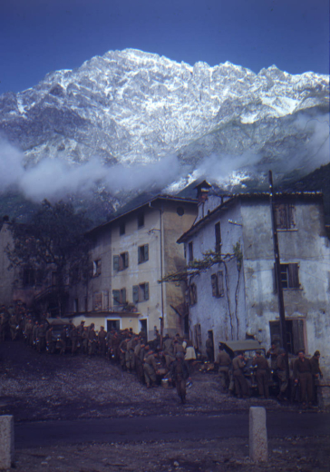 Shows a long line of more than thirty Tenth Mountain Division soldiers waiting for food. The chow-line snakes up a steep street in an Italian town; a few men are eating around a covered jeep.  Above the town is a rugged mountain covered with snow; clouds hover over the roofs.