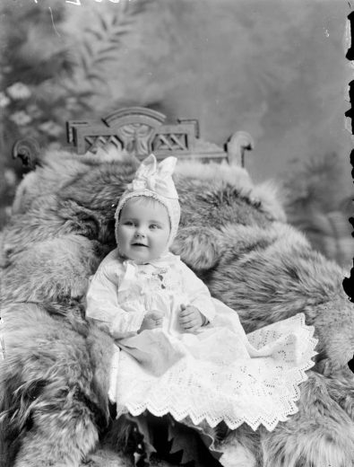 Studio portrait of a baby girl. She poses on a chair covered with a fur blanket and wears a dress and a bonnet decorated with ribbon.