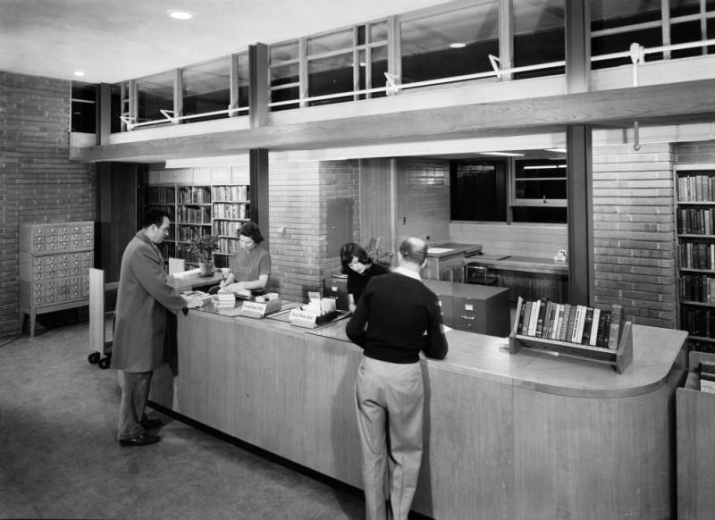 Interior view of the Ross-Broadway Branch of the Denver Public Library in the Speer neighborhood of Denver, Colorado; men wait at a counter and women work. Decor includes card catalogs, bookshelves, and monitor windows.
