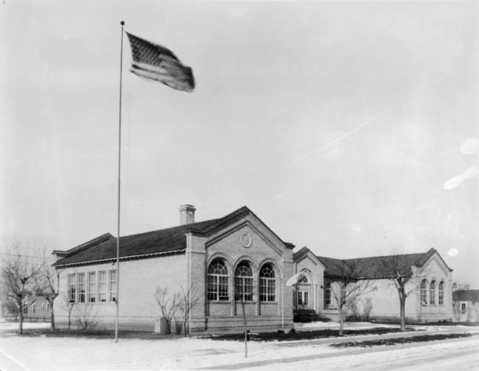 View of Elyria School at 4725 High Street in the Elyria-Swansea neighborhood of Denver, Colorado; the brick building has arched windows and a tile roof. A United States flag waves on a pole.
