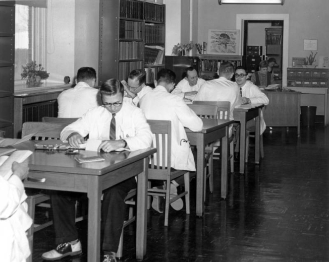 A group of doctors or possibly medical students sit at tables in a library, a branch of Denver Public Library, at Denver General Hospital in Denver, Colorado. The men wear white jackets and have books open in front of them. One man wears saddle shoes. Books are arranged on shelves along the walls. Two women librarians sit at desks in the distance.
