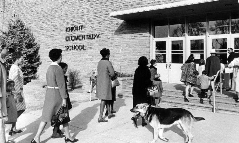 African-American (Black) and other women and children enter Knight Elementary School (later Knight Fundamental Academy) at 3245 East Exposition Avenue in the Belcaro neighborhood of Denver, Colorado. An African-American man turns and smiles at the women and children as he opens the door. A German Shepherd dog watches the people enter the school.