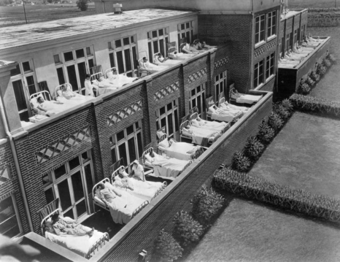 Tuberculosis patients lie in beds on the porch of a building at the Jewish Consumptive Relief Society (J.C.R.S.) sanatorium, 1600 Pierce Street, Lakewood, Colorado. The men lie in rows outside their rooms, and nurses attend to some patients.