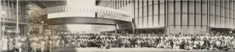 Panoramic view of African American (Black) women, members of the National Association of Colored Women's Clubs Inc. and the N.A.G.C., men, boys, and girls pose on Court Place in downtown Denver, Colorado. A covered pedestrian bridge is over the street. Signs read: "Creative Jew[elry]," "Denver Hilton," and "May D & F."