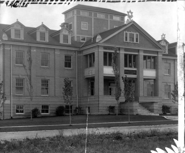 Exterior view of Beth Israel Hospital on 16th (Sixteenth) Avenue and Lowell Boulevard in the West Colfax neighborhood of Denver, Colorado. The brick institutional structure features a row of dormers and decorative Star of David finials. Lettering on the two-tiered portico reads: "Leopold H. Guildman Building" and "Beth Israel Hospital."
