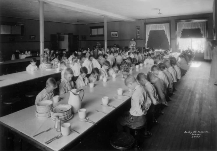 Boys sit at long tables in prayer before mealtime at the Denver Orphans' Home (later the Denver Children's Home) at 1501 Albion Street in the South Park Hill neighborhood of Denver, Colorado. Shows a china mug, knife, fork and spoon at each place setting. A flowering plant is on each table.