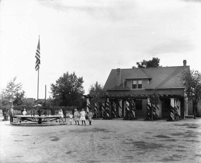 Children pose next to the merry-go-round and flagpole at the Elyria Community Center in Elyria Park, 4809 High Street in the Elyria Swansea neighborhood of Denver, Colorado. Shows a two story brick building with a dormer and a stone column pergola.
