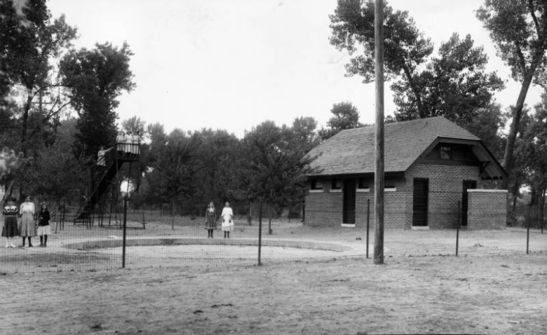 Girls stand at the top of a slide and around a circular sandbox at probably Argo Park at 47th (Forty-seventh) and Logan Streets in the Globeville neighborhood of Denver, Colorado. Shows a brick structure with bathrooms and a Dutch hip roof.