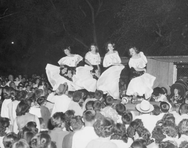 The Auraria Casa Mayan dancers entertain at a 16th of September (Diez y Seis de Septiembre) Mexican Independence Day celebration in Curtis Park in the Five Points neighborhood, Denver, Colorado. Dancers include Magdalena Gallegos and Alice Montoya. The young women wear matching dresses with full skirts and sleeves, scoop necks and dark shawls wrapped around their waists. Men, women and children are crowded around the stage.