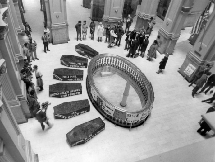 Interior view of the Colorado State Capitol in Denver, Colorado during a political demonstration. People, fake coffins, and picket signs surround the mezzanine railing. Signs on coffins read: "Justice Miss Farm Labor," "Mr. Education," "Police Respect Law and Order," "Mr. & Mrs. Housing," "Consumer Protection," and "Mrs. Welfare." A picket sign reads: "Justice is Dead."