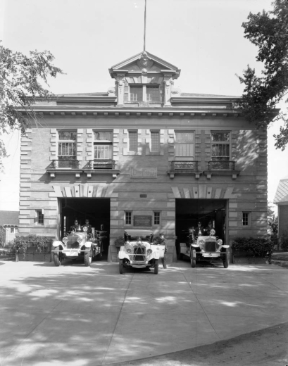 View of Denver Fire Department Station No. 15 at 1080 Clayton Street in the Congress Park neighborhood of Denver, Colorado. The stone building has quoins, voussoirs, and a gable. Firemen pose in fire trucks.