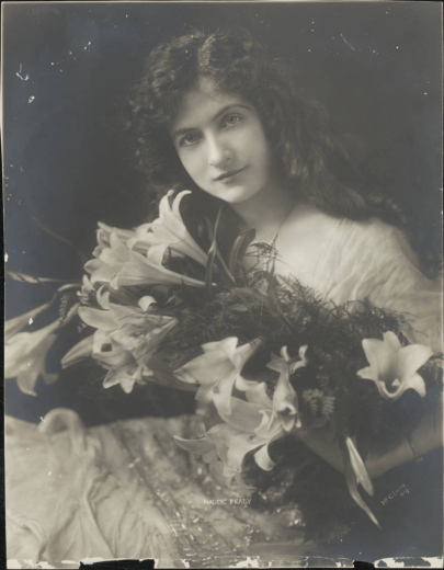 Maude Fealy poses for a studio bust portrait, possibly for the role of "Filiberta" in "The Cardinal." She has long, wavy hair; wears a bracelet, a pearl necklace, and a dress with gathered, sheer fabric and beadwork; and holds a bouquet of lilies.