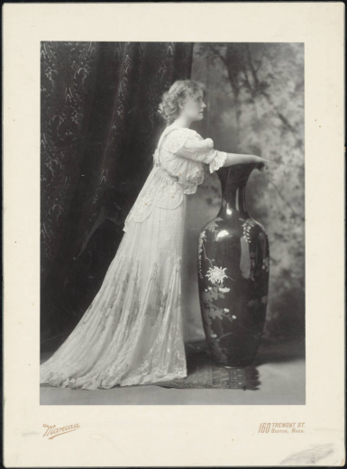 Studio profile portrait of Antoinette Perry, an actress, who stands and poses with her arms extended over a large, ceramic vase with painted flowers. She wears a long two piece lace dress with a slim skirt and pleats midway down the thigh. Her blouse has a scalloped and tapered waist, shoulder ruffles, and short, gathered sleeves. A lace sash connects at the back under probably a pearl charm detail. Antoinette Perry has wavy hair pulled back with ringlets.