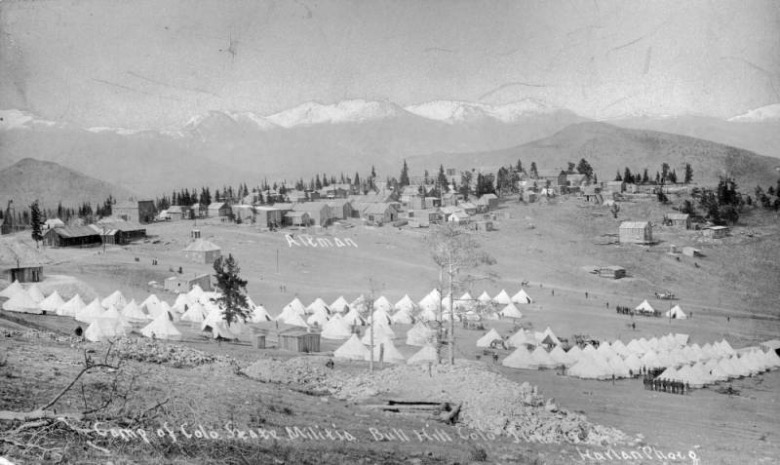 Panoramic view, probably during a mining labor strike, of frame buildings in the town of Altman (Teller County), Colorado and tents in rows at a military encampment in a nearby meadow. A flagpole with an American flag is near tents, and soldiers in uniform stand in rows. Frame commercial buildings and houses and possibly Western Federation of Miners buildings are in Altman. Shows a portion of Pikes Peak amid snowy mountains.