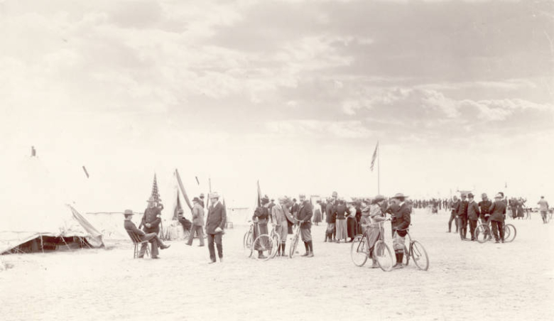First Colorado Regiment soldiers, officers, and visitors, men and women, stand by bicycles, tents, and United States flags. Outfits include uniforms, gaiters, a puff shouldered blouse, and a straw hat.
