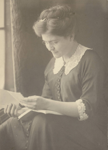 Josephine A. Roche reads a folded paper and has a book in her lap. Her hair is upswept, and her blouse has lace trim.