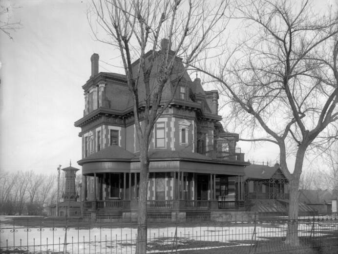 The large, three-story home of Marcus Mills "Brick" Pomeroy stands at 2949 West 37th (Thirty-Seventh) Street at the corner of Federal Boulevard in Denver, Colorado. Designed by Pomeroy, the home included an art gallery and a speaker's platform. Two trees with bare branches stand near a wrought iron fence that borders the property in the foreground.