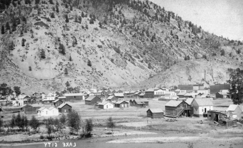 Several log cabins and wood frame buildings line dirt streets at the base of a mountain in the town of Lake City in Hinsdale County, Colorado. A small creek runs through the foreground.