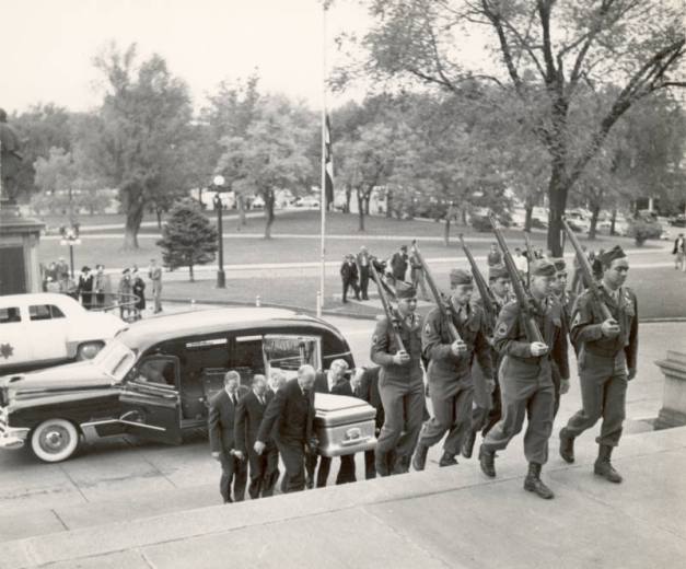 Pallbearers carry the coffin of Ralph L. Carr up steps at the Colorado State Capitol in Denver, Colorado. A military honor guard with rifles leads the way; a hearse is at the curb, and people watch from capitol grounds. A Colorado flag flies at half-mast.
