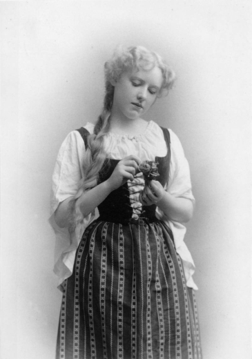 Studio portrait of Denver actress Antoinette Perry. She wears a white blouse, a laced, velvet bodice and a skirt with embroidered stripes. She wears her hair in a long braid and holds a rose.