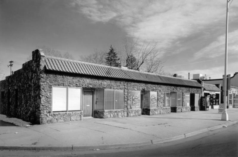 View of storefronts at 3938-42 Federal Boulevard in the Sunnyside Neighborhood of Denver, Colorado; features include moss rock cladding and a terra cotta tile pent roof. Show windows and lettering reads: "Insurance Able Specialists," and "Remedio Juarez."  A vending machine reads: "Coca Cola."