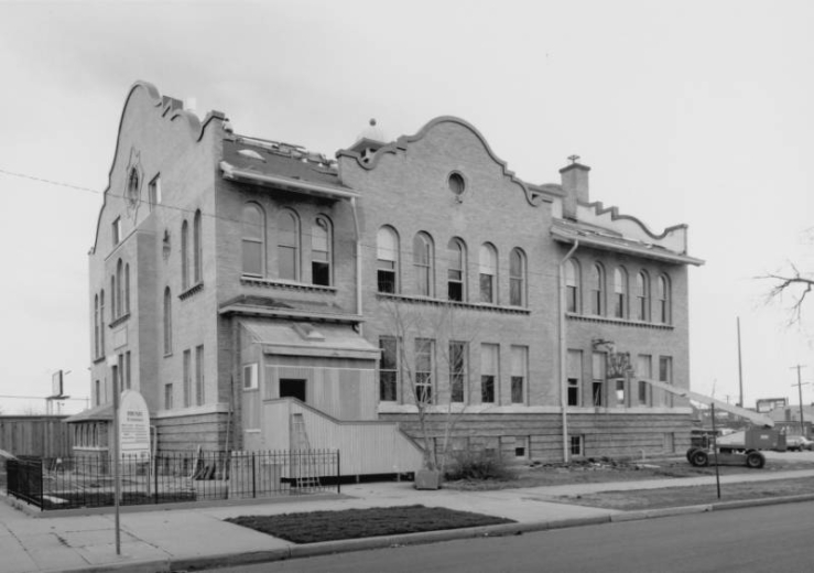 View of the former Byers Elementary School at 104 West Byers Place in the Baker Neighborhood of Denver, Colorado. The brick building converted to condominiums has Mission Style pediments, arched and round windows, a round balcony, and steps. A man works on the building from a light duty telescoping crane.