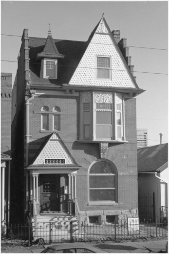 View of a three story brick house at 2418 Stout Street in the Five Points neighborhood of Denver, Colorado. The house has eclectic architectural elements that include arched windows with voussoirs, bargeboard gables, corbiestep sides, plaster relief details, and a dormer. A sign in the yard reads: "Offered by Marsh & Associates, 628-5474, For Rent."