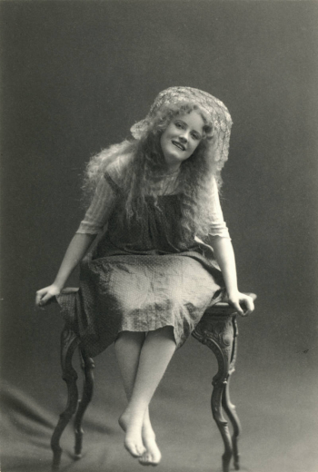 Studio portrait of Denver actress Antoinette Perry. She wears a woven straw hat with her hair down, a pinafore with a pleated bodice, and a striped blouse trimmed with lace. She is barefoot. She smiles, and poses on a cast iron table with her legs crossed at the ankles.