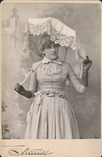 Studio portrait of Mrs. Leonel Ross Anthony O'Brien holding an umbrella with lace trim. She wears leather gloves and a dress with a pleated waist line, buttons, and lace collar.