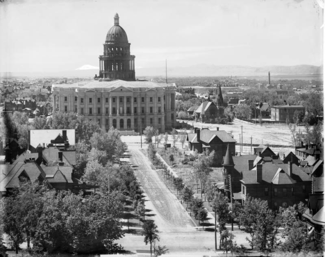 View of the Colorado State Capitol Building in Denver, Colorado from the Central Presbyterian Church tower on Sherman Street. The dome of the capitol building is under construction and scaffolding braces are in place inside the tower. The First United Presbyterian Church is nearby. Buildings and a smokestack of Denver General Hospital are in the distance.