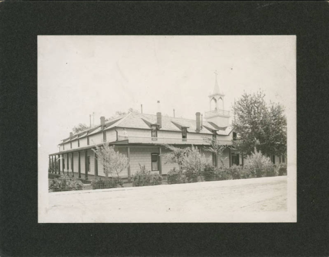 View of the Sisters of Loretto Academy and convent in Las Vegas (San Miguel County), New Mexico. Shows an adobe building with siding and Territorial style architectural elements. The building has a portal, a balcony, dormers, chimneys, and a wooden steeple topped with a cross. A woman poses on the balcony. Trees and bushes are in the yard.