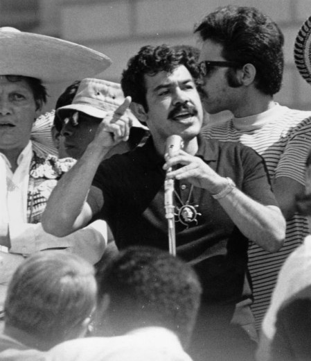 Mexican American Rodolpho "Corky" Gonzales, Mexican American activist and founder of the Crusade for Justice, addresses a crowd in probably Denver, Colorado. He holds a microphone and wears a pendant. A man in a large straw sombrero stands nearby.