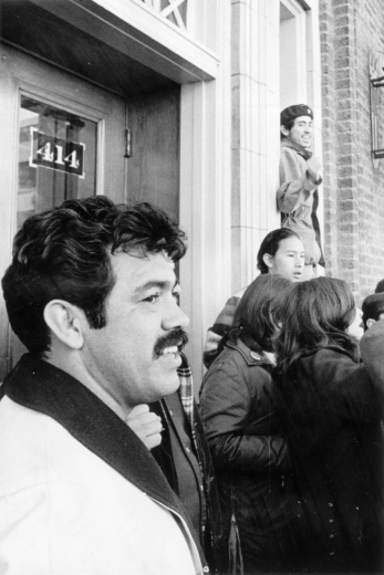 Rodolpho "Corky" Gonzales, Mexican American political activist and founder of the Crusade for Justice, stands with young Mexican American men and women near a doorway in Denver, Colorado. A man in a beret stands close by and raises his fist.