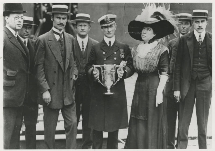 Margaret Tobin (Molly) Brown and Captain A. H. Rostron of Cunard Linespose with an engraved silver trophy cup. Five men in suits, bowler or straw hats (one with a pince-nez) look on; the ceremony commemorates Brown's rescue from the Titanic.