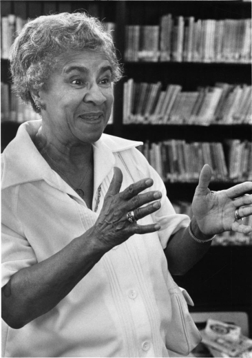 Photograph of Marie Greenwood speaking at the Read Aloud Volunteer party. She wears a light colored blouse and is standing in front of a bookshelf.